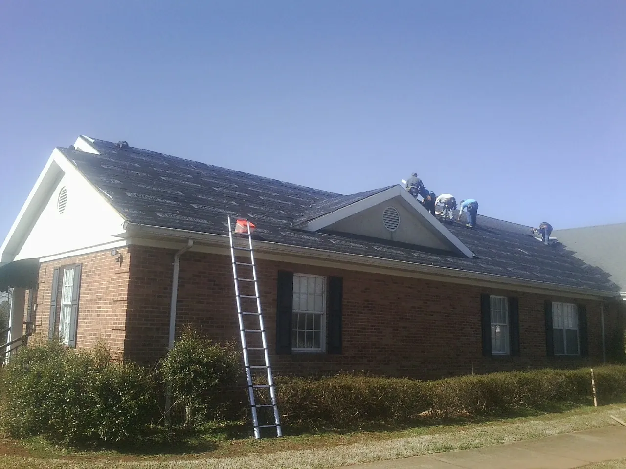 Crew tearing shingles off a brick ranch roof with a ladder set — Emergency Roof Repair work by our crew in Oak Hill