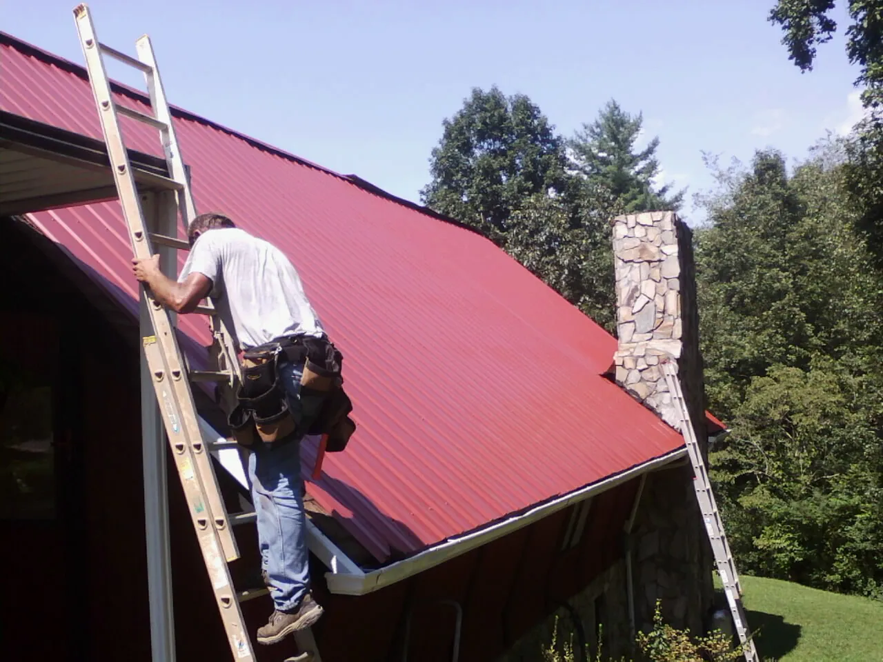 Roofer on a ladder beside a red metal roof and stone chimney — Soffit & Fascia Repair by Roofing Brothers in Oak Hill